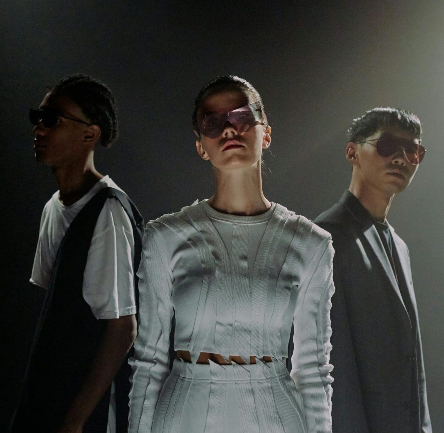 Three models in stylish attire and sunglasses standing in a dimly lit studio space with a dramatic mood.