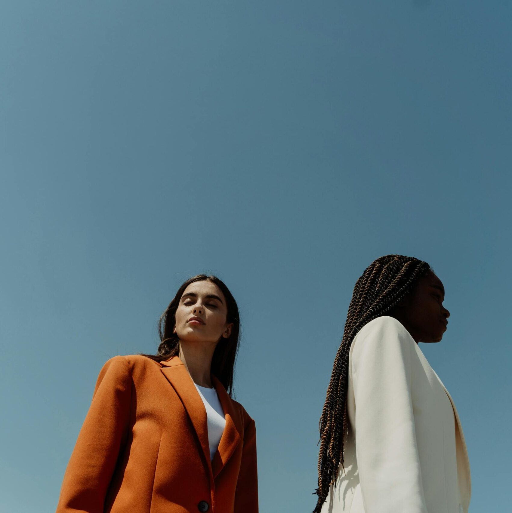 Two stylish women in corporate attire against a blue sky.
